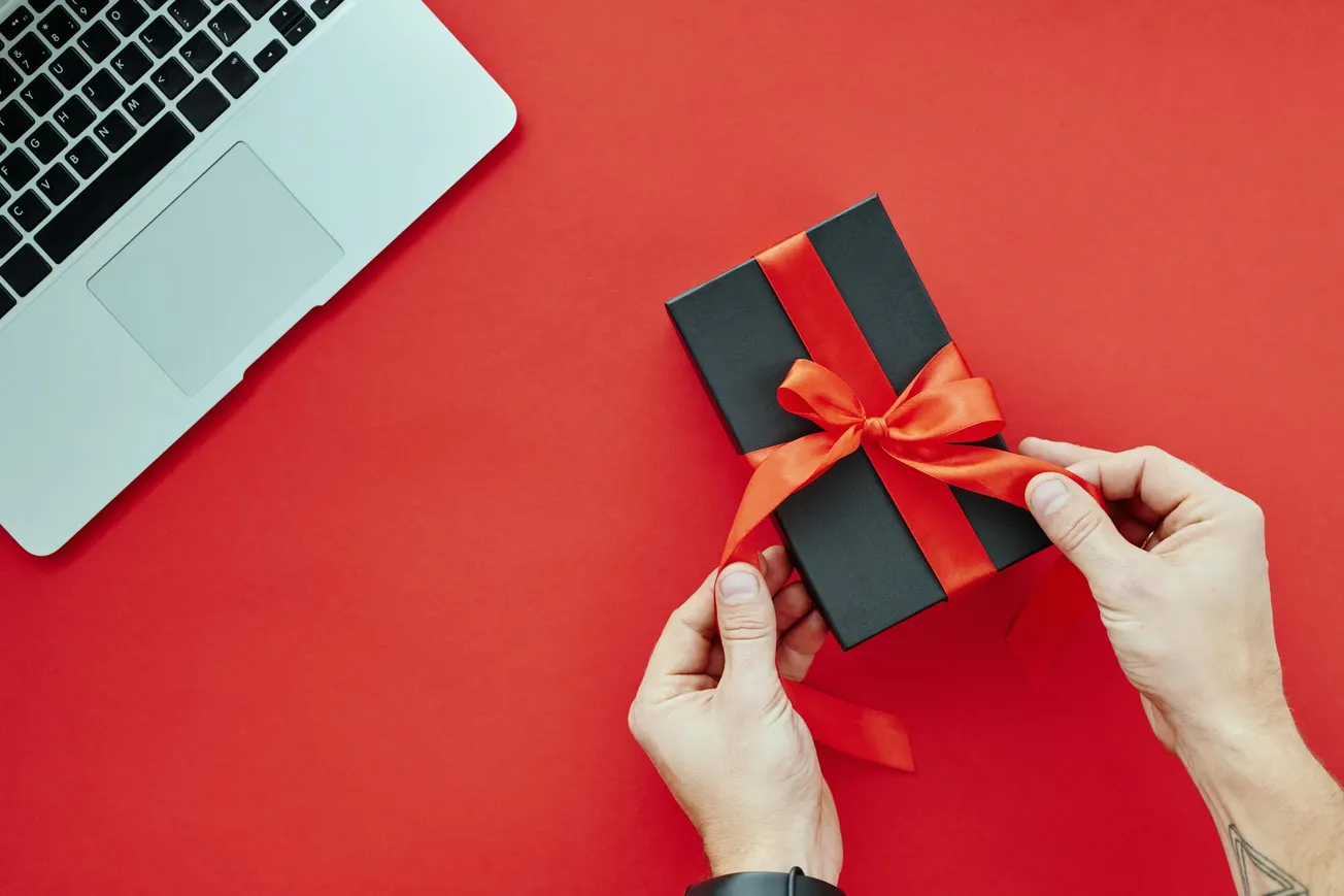 Hands untying a red ribbon on a black gift box, set on a vibrant red surface next to a silver laptop. The scene conveys anticipation and excitement.