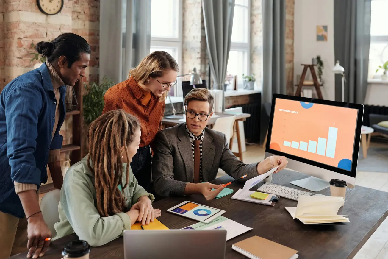 A diverse group of four people engaged in a collaborative meeting around a table with charts. A computer screen shows a bar graph, suggesting data analysis.