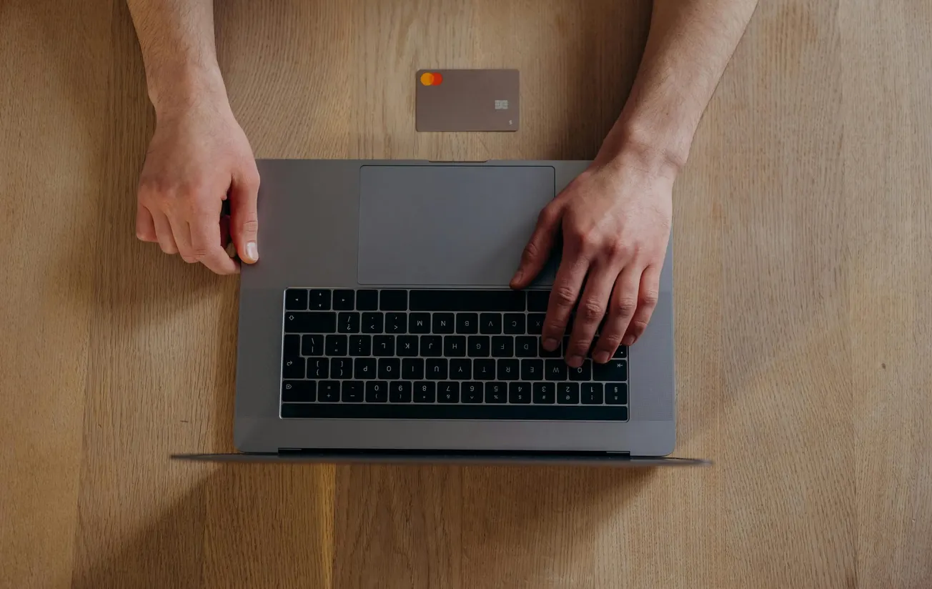 Two hands on a laptop keyboard beside a credit card on a wooden table, conveying a theme of online shopping or digital payment.