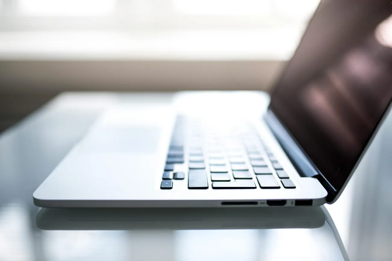 A laptop sits open on a reflective white table, angled with the keys and screen partially visible. Sunlight streams through a window in the background.