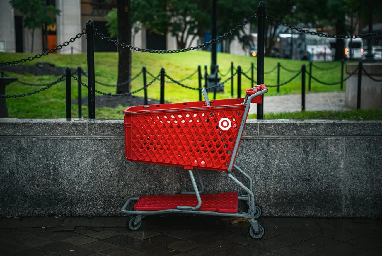 A solitary red shopping cart is parked against a gray stone wall. It stands near a black chain-link fence. The background shows green grass and trees.