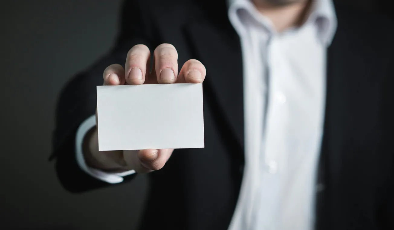 A person in a suit holds a blank white business card toward the camera against a dark background, conveying a professional and formal tone.