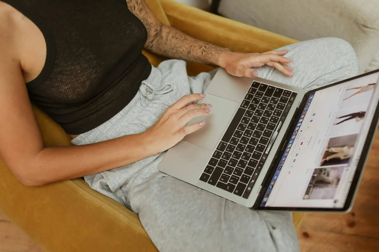 A person in a black top and gray sweatpants sits in a yellow chair, browsing fashion images on a laptop. The scene is relaxed and casual.