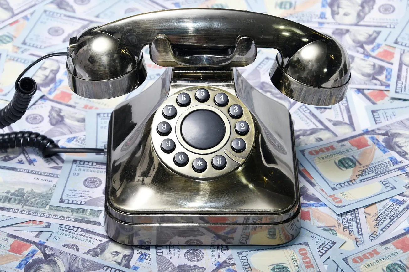 A shiny, vintage rotary phone is placed on a spread of one hundred dollar bills. The image conveys a sense of wealth and retro communication.