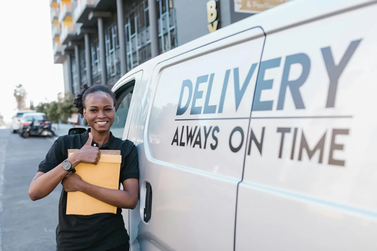 Smiling delivery worker holding an envelope stands beside a van labeled "DELIVERY ALWAYS ON TIME" on an urban street, conveying reliability.