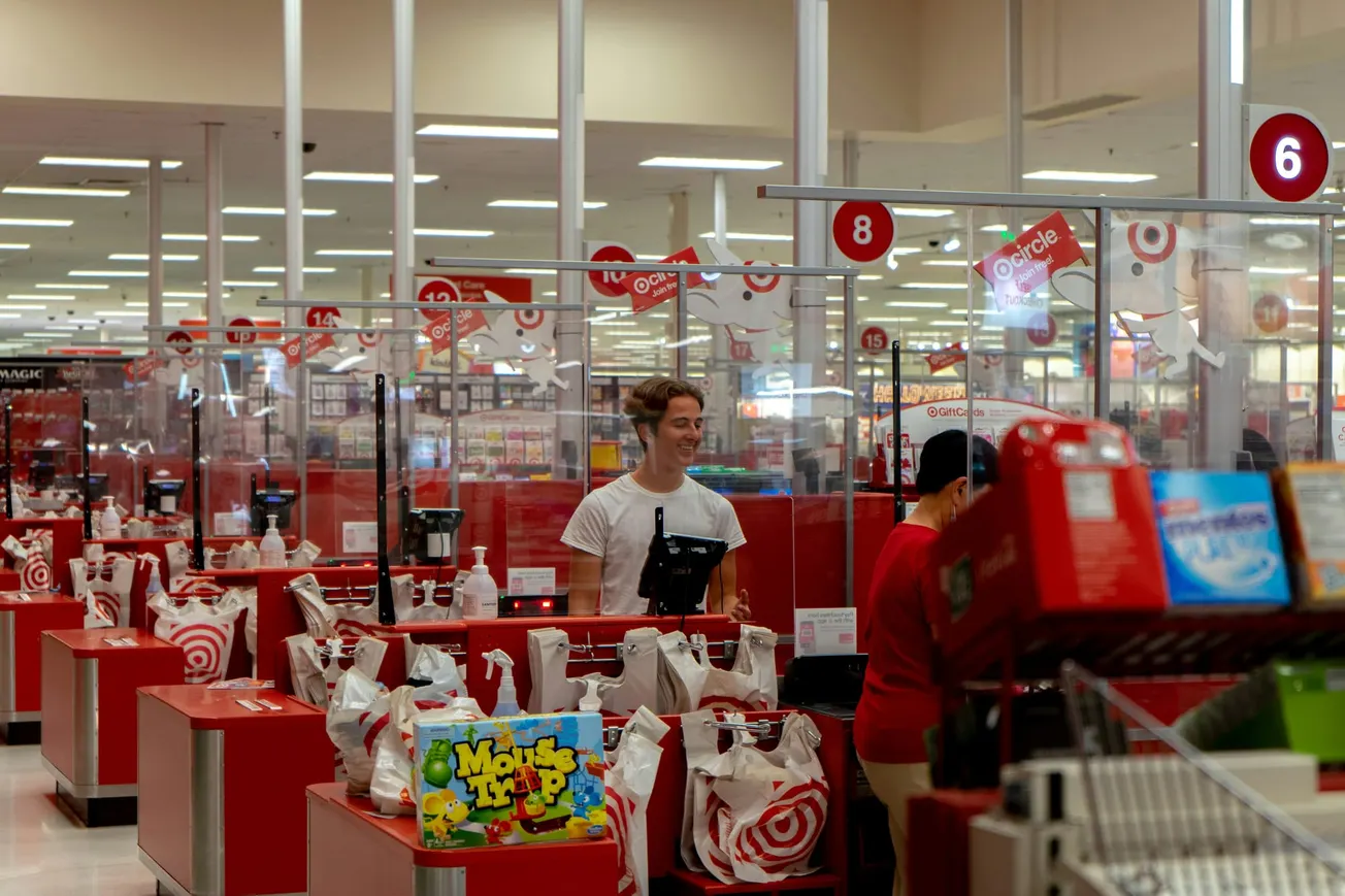 A busy retail store checkout area with red counters and clear barriers. Customers, cashiers, and shopping bags with logos are visible, creating a lively atmosphere.