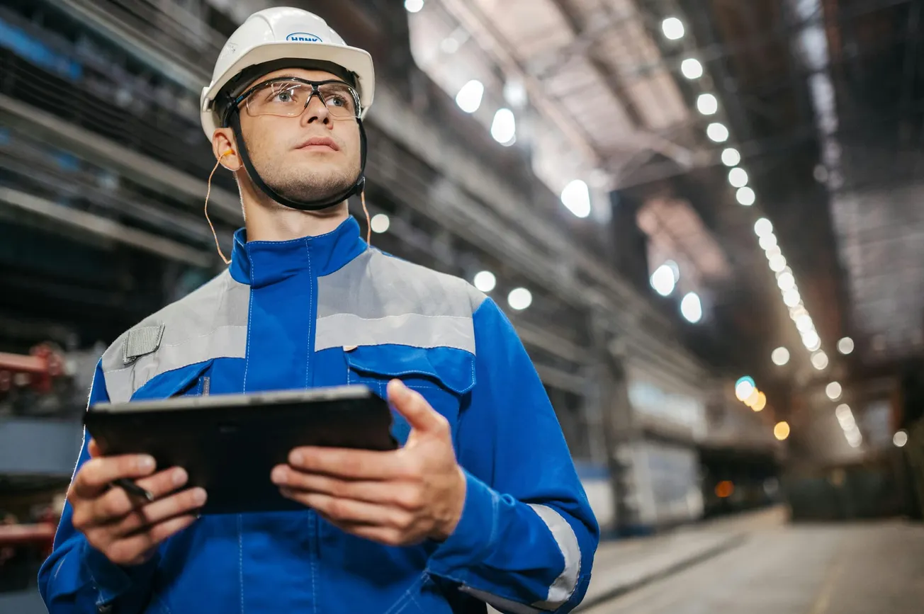 A worker in a blue uniform and protective gear holds a tablet, standing in a large, well-lit industrial facility. The scene conveys focus and professionalism.
