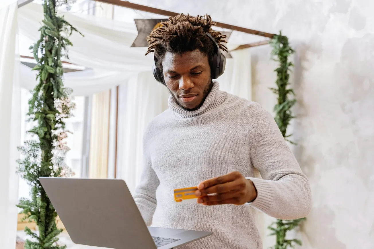 A man in a cozy sweater with headphones holds a credit card, using a laptop. Behind him, festive green garlands convey a holiday atmosphere.