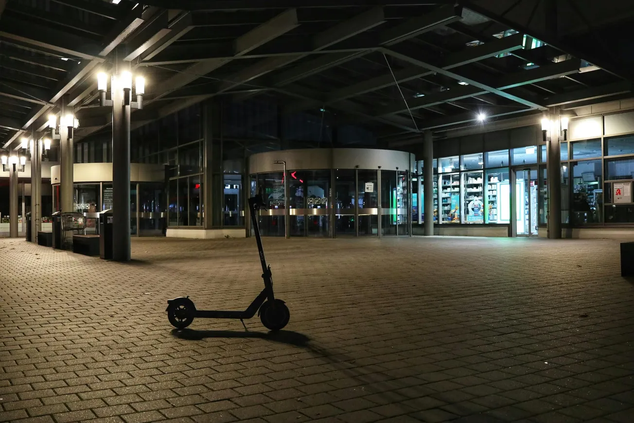 Electric scooter stands alone on a dimly lit, empty plaza at night, surrounded by large glass windows and overhead lights casting soft glows.