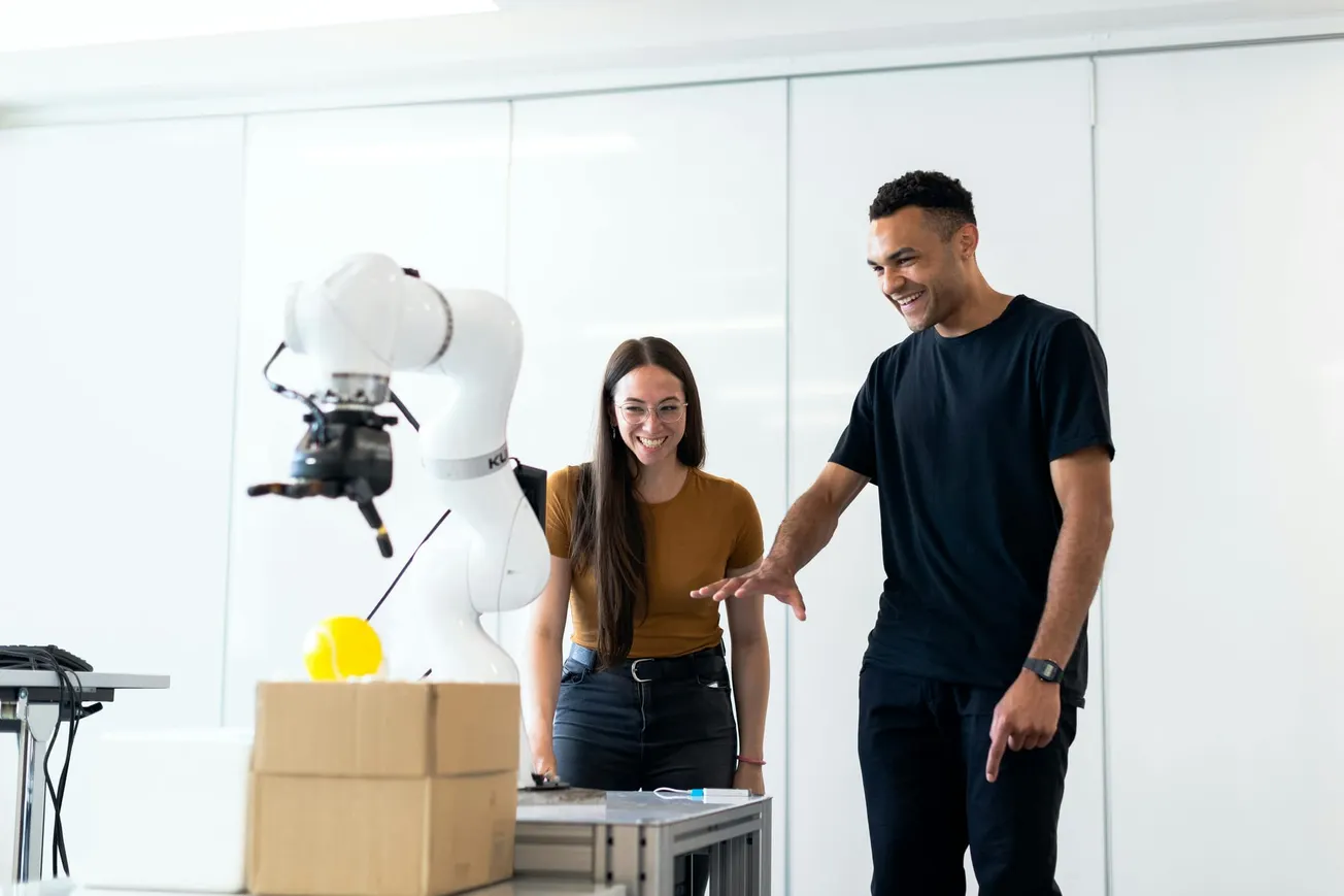 Two people joyfully interact with a robotic arm in a bright, modern lab. The arm holds a yellow object, and cardboard boxes are visible on a desk nearby.