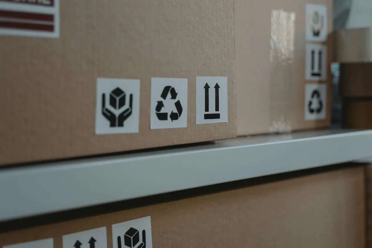 Brown cardboard boxes on a shelf, featuring handling symbols: fragile, recycling, and arrows indicating correct upright position. Represents packaging and shipping.
