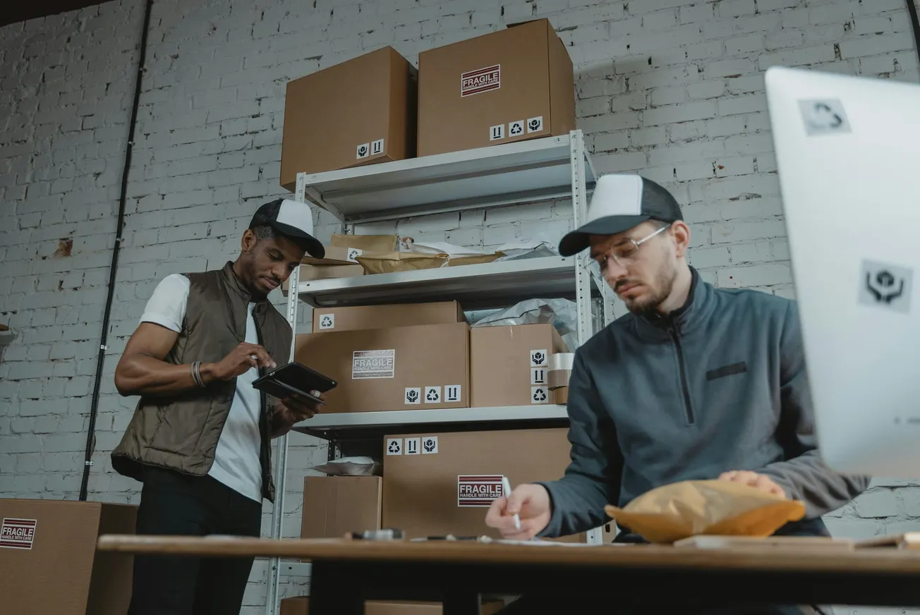 Two workers in a warehouse are organizing packages. One inspects a tablet, the other writes notes. Cardboard boxes labeled "Fragile" are stacked behind.