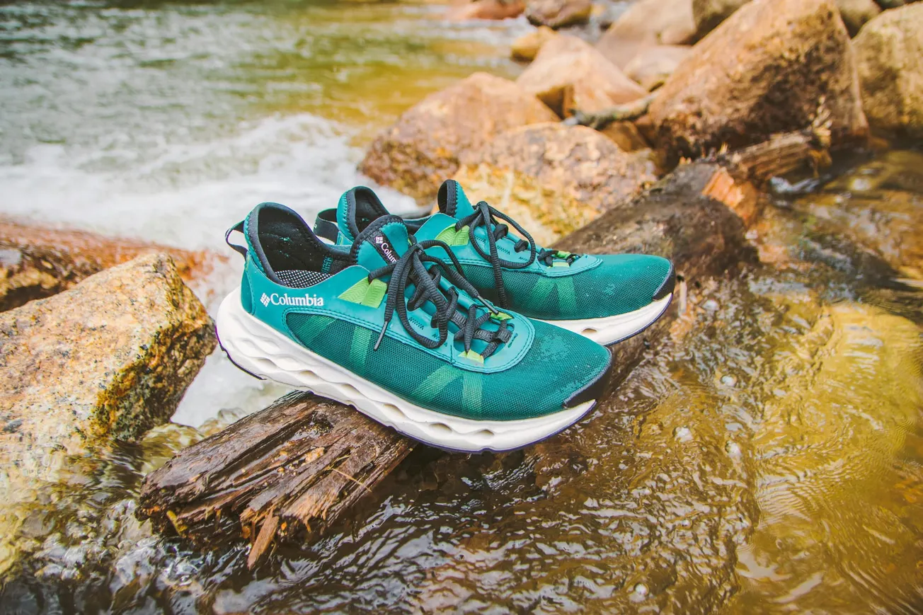 Teal athletic shoes with black laces rest on a log in a flowing river. Surrounding rocks and water create a natural, adventurous atmosphere.