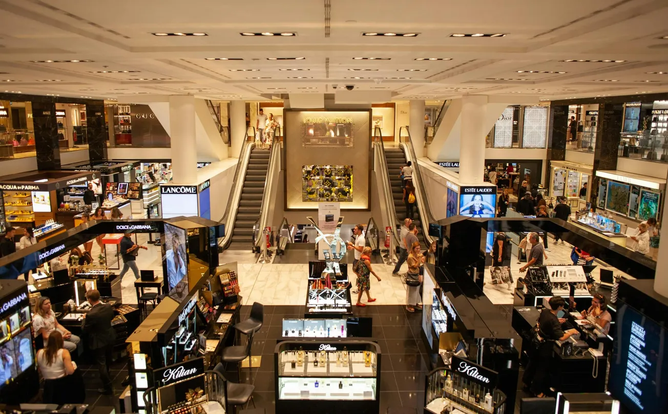A bustling department store interior features makeup displays, shoppers browsing, and escalators in the center, creating a lively and vibrant atmosphere.