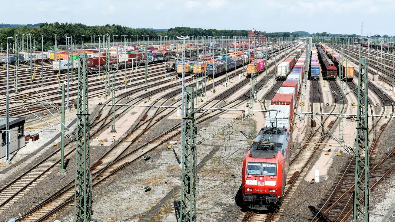 A vast railway freight yard with numerous tracks filled with colorful shipping containers. A red locomotive moves forward, conveying busy activity and industrial efficiency.