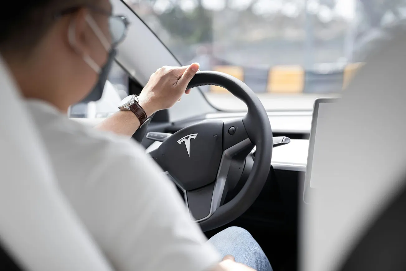 A person wearing a mask is driving a Tesla, focusing on the road. The Tesla logo is visible on the steering wheel, conveying a modern and safe atmosphere.