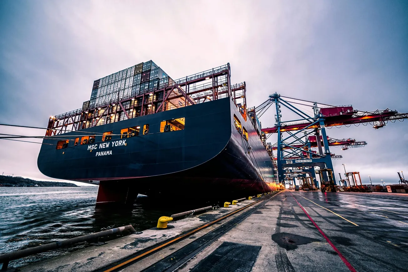 A large cargo ship, "MSC New York," is docked at a port with towering cranes nearby. The scene is captured at dusk, conveying a bustling industrial atmosphere.
