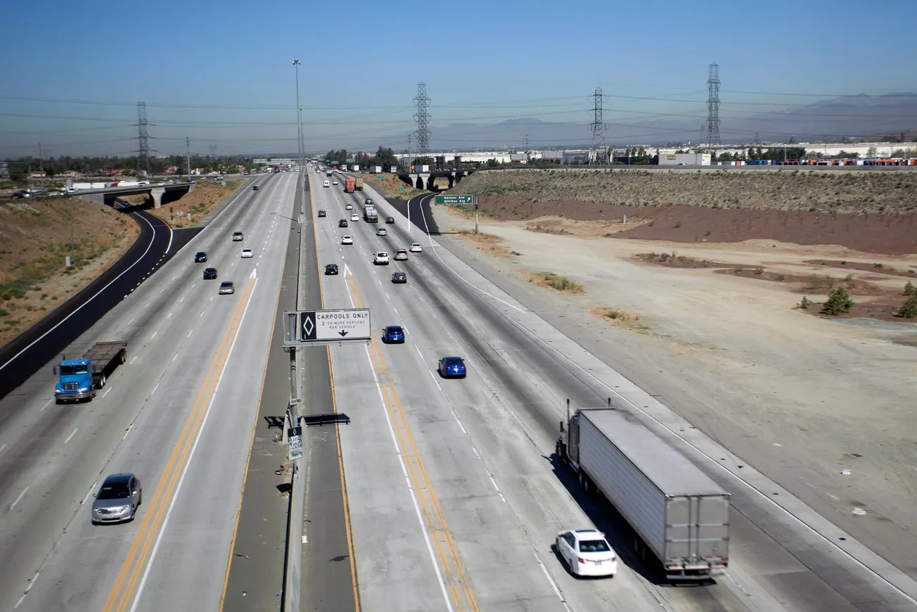 Wide highway in daylight, sparse traffic with trucks and cars, bordered by desert landscape. Clear sky, industrial buildings in the distance. Calm atmosphere.