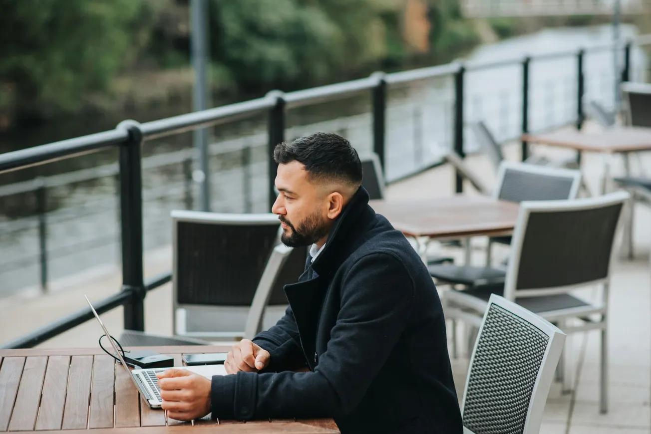 A man in a dark coat works on a laptop at an outdoor café table by a river. The setting is calm and scenic, with empty tables and greenery in the background.