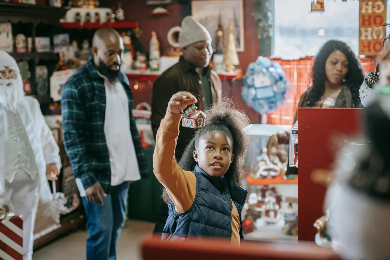 A young girl in a store holds up a festive ornament, surrounded by family. The shop is decorated for the holidays, creating a warm, joyful atmosphere.