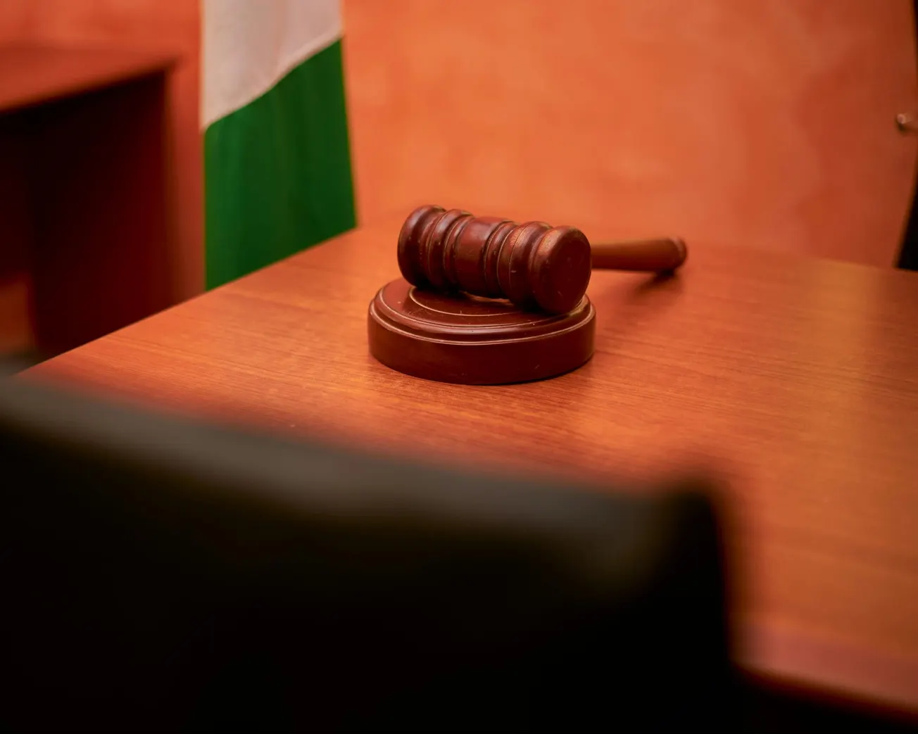 A wooden gavel rests on a courtroom desk with a blurred green and white flag in the background, conveying a sense of authority and justice.