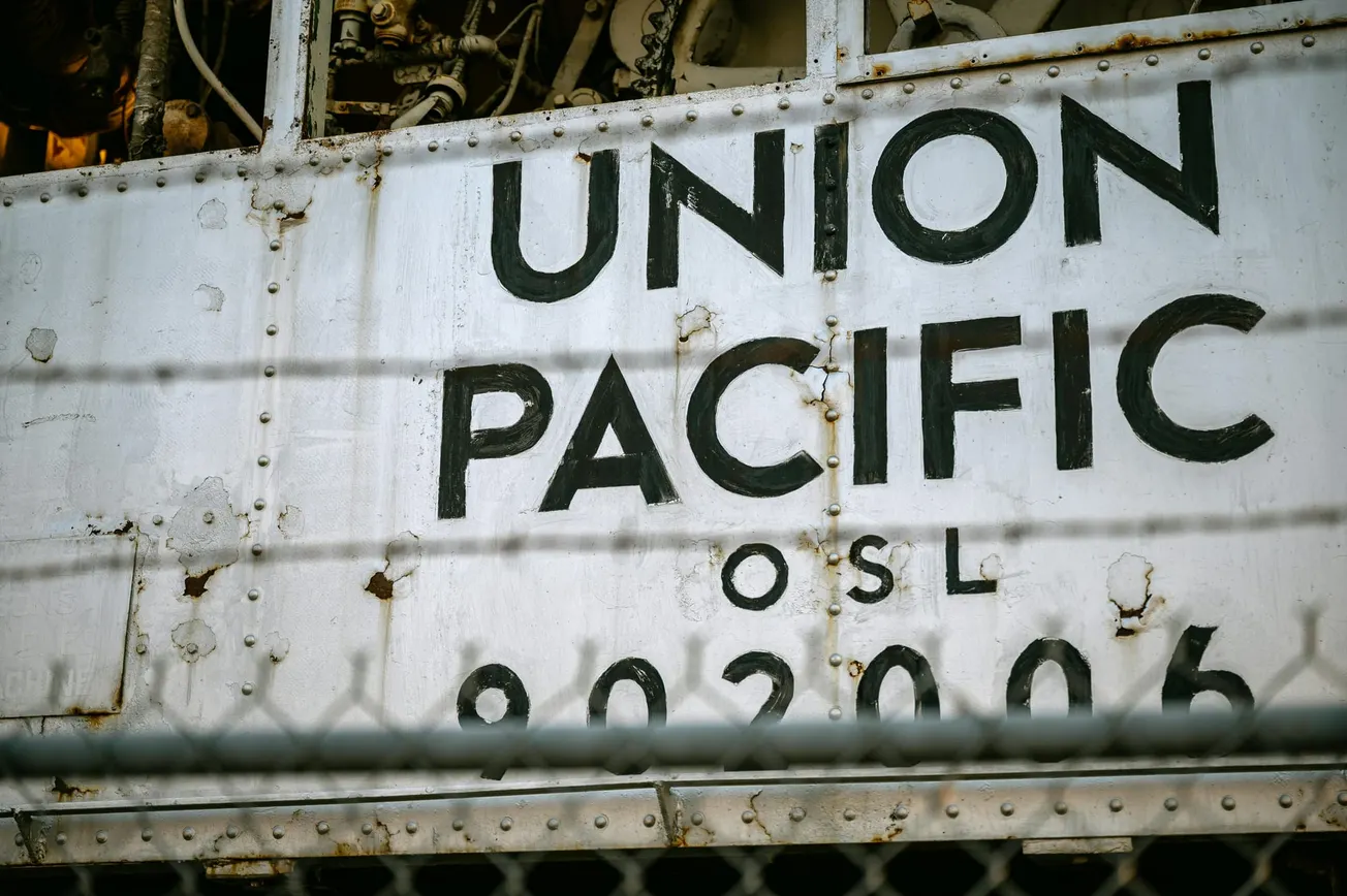 Close-up of a weathered train car with "Union Pacific OSL 902006" in bold black letters. A chain-link fence and barbed wire are visible in the foreground.