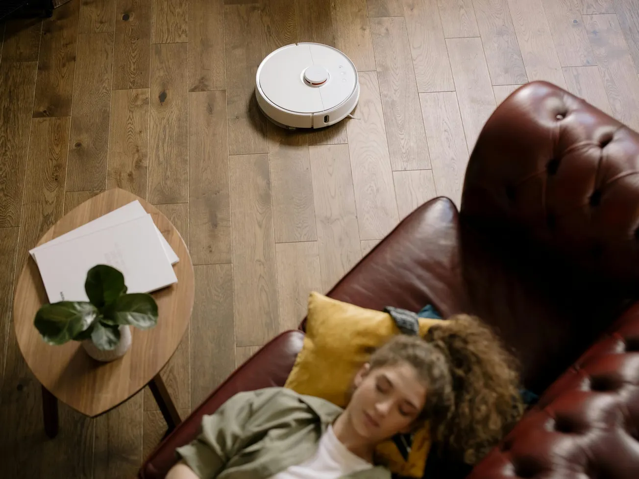 A person with curly hair sleeps on a brown sofa, while a robotic vacuum cleans the wooden floor. A plant and papers rest on a side table nearby.