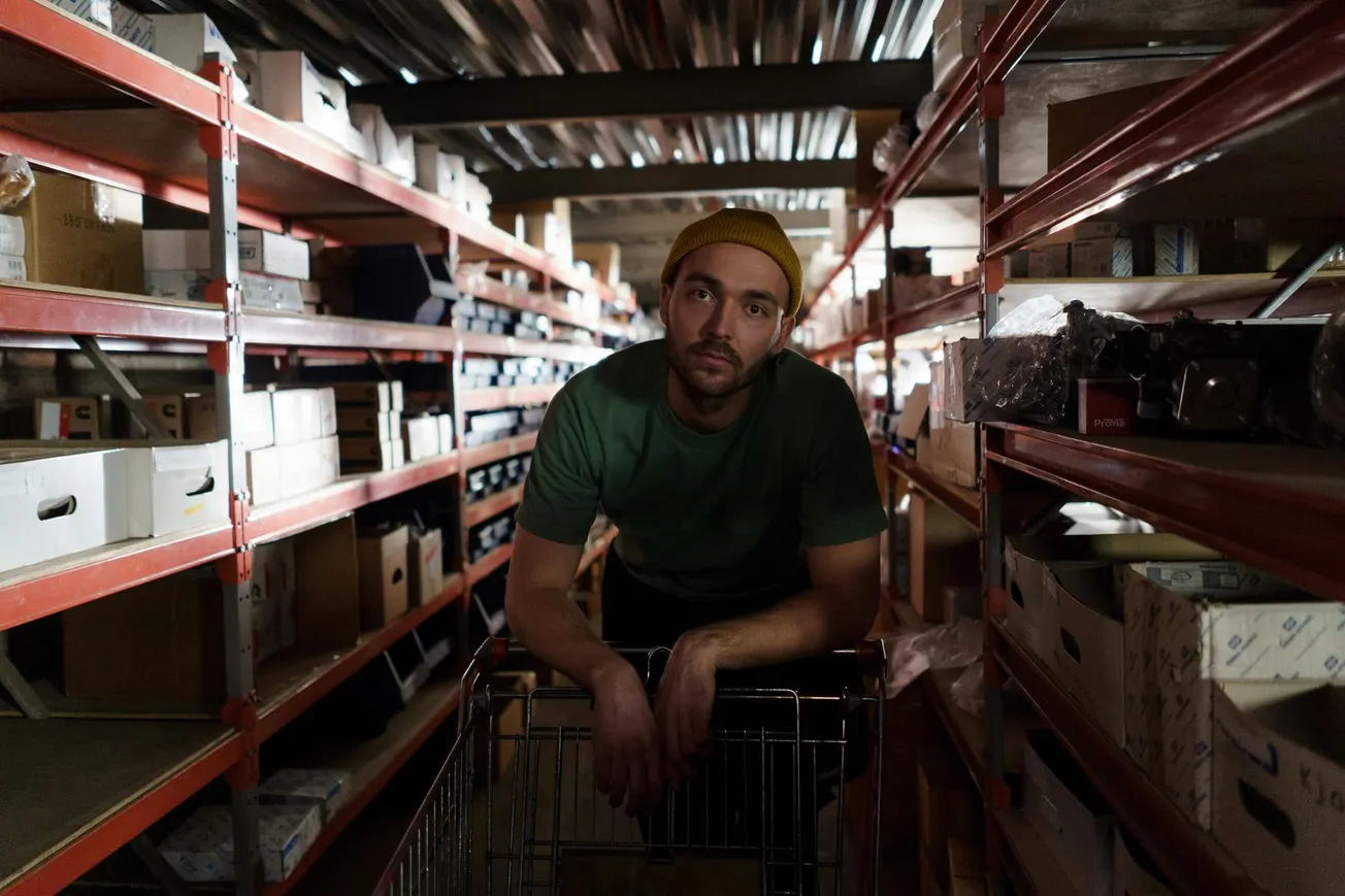 Man in green shirt and yellow beanie leans on shopping cart in dimly lit warehouse aisle. Shelves with boxes line both sides, creating a focused, contemplative mood.