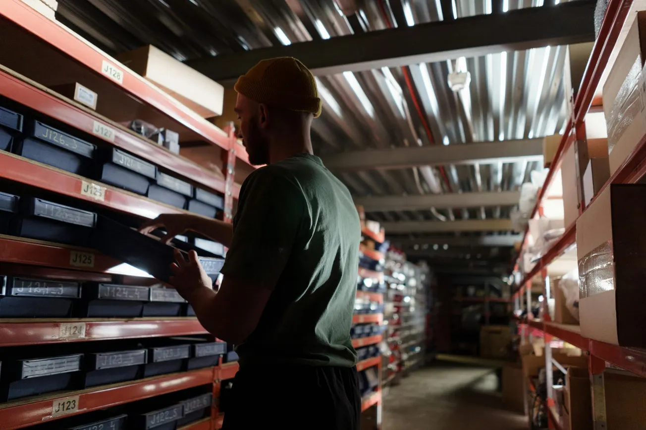 A person in a dimly lit warehouse looks at stacked boxes on metal shelves, creating a focused and industrious atmosphere.