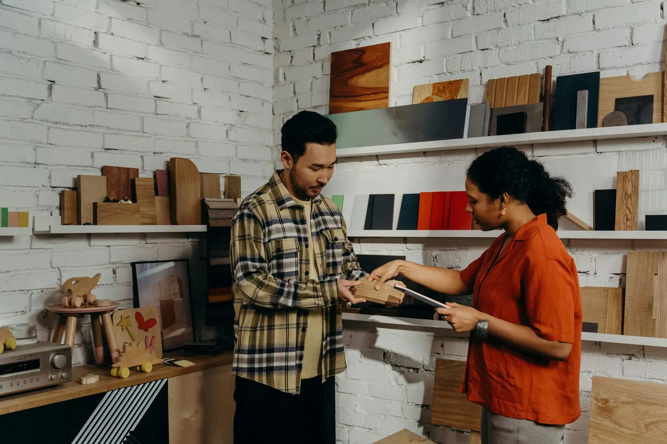 A man and woman are discussing wood samples in a design studio with shelves full of materials. The setting conveys creativity and collaboration.