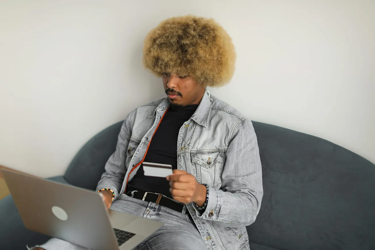 A person with curly hair sits on a couch, wearing a denim jacket, and focused on a laptop while holding a credit card.