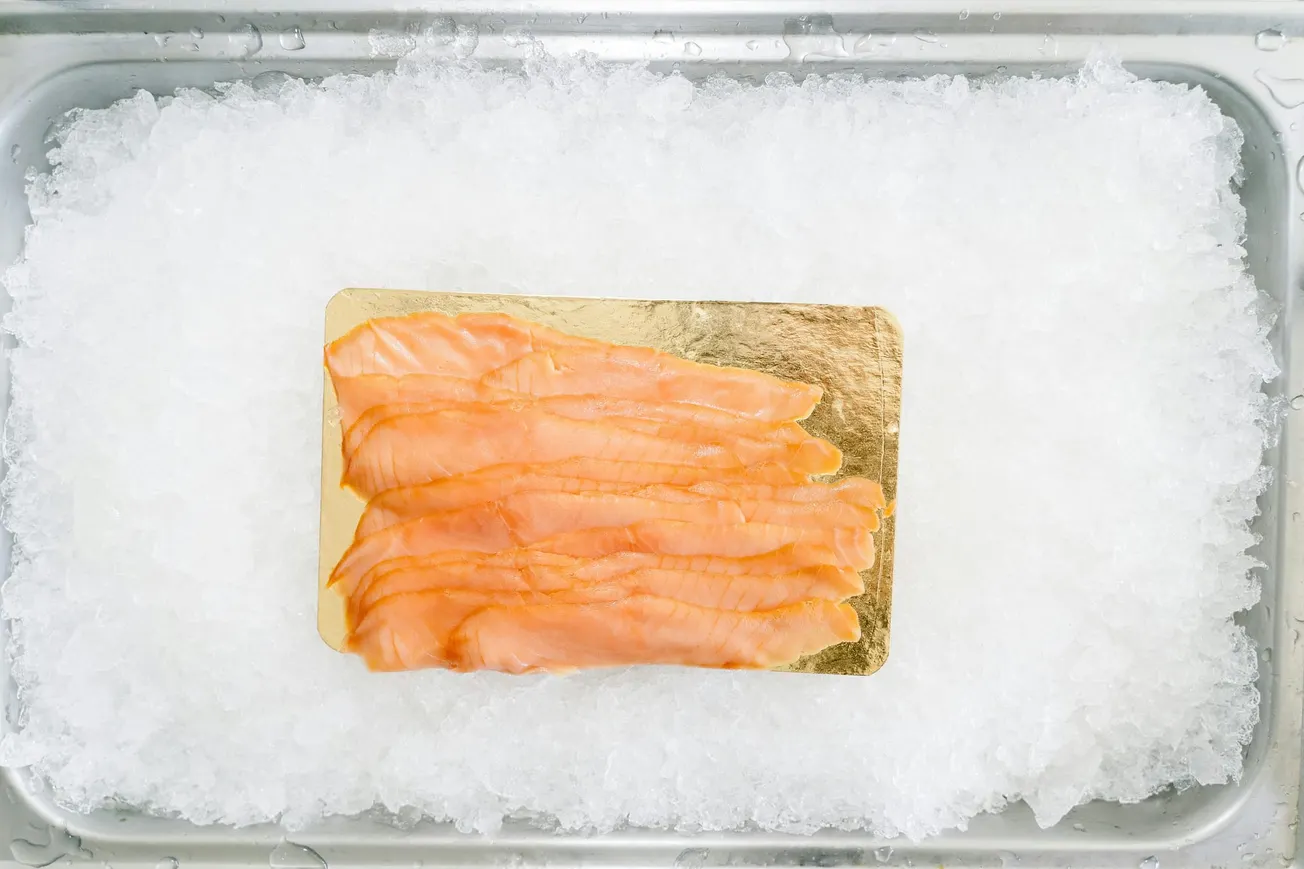 Slices of smoked salmon arranged on a gold-colored board resting on a bed of crushed ice in a metal tray, creating a fresh, chilled presentation.