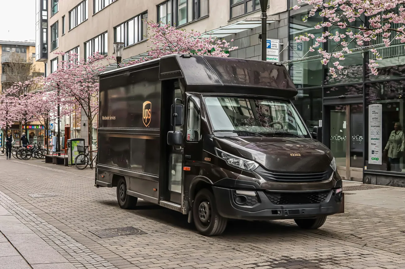 A delivery truck parked on a street lined with blooming cherry blossoms. Modern buildings in the background create a serene, urban spring scene.