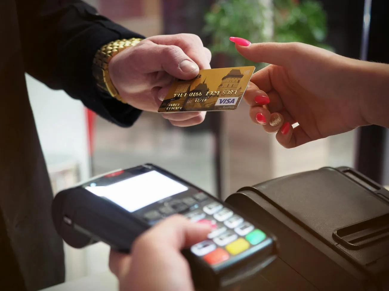A person hands a credit card to another individual over a payment terminal. The scene conveys a transaction, emphasizing convenience and technology.
