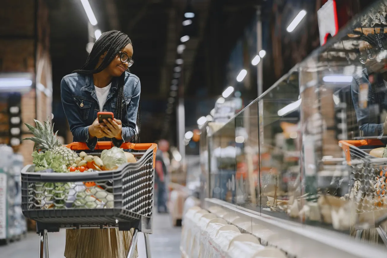 A smiling woman with braided hair, in a denim jacket, leans on a shopping cart filled with fruits and vegetables, using her phone in a grocery store aisle.