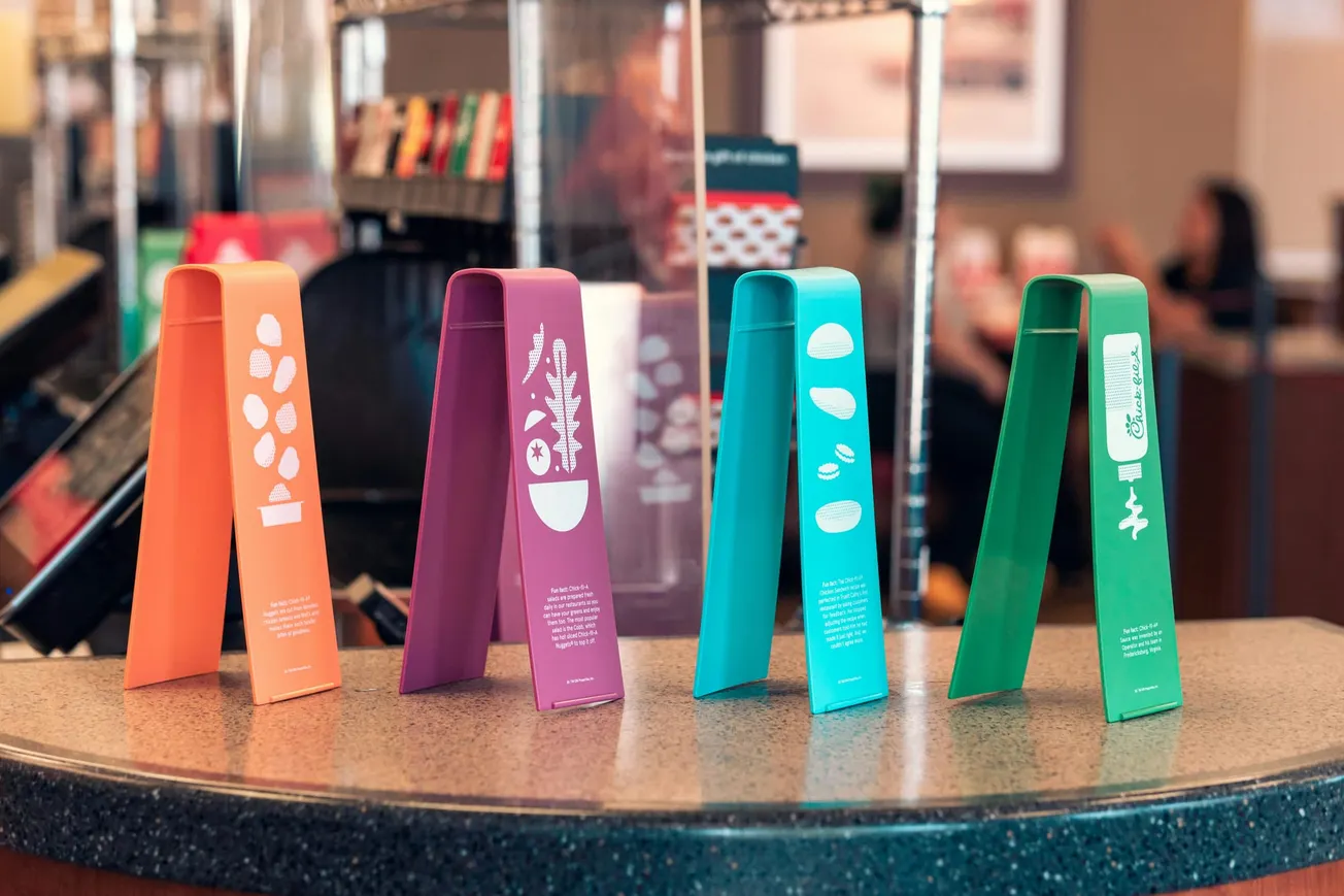 Four colorful, tent-shaped signs on a counter display food illustrations and text. The vibrant orange, purple, teal, and green signs add a lively touch.