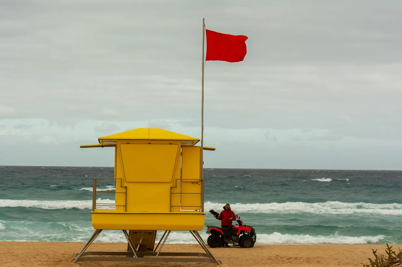 Yellow lifeguard tower on a sandy beach, with a red flag indicating danger. A person on a red ATV is nearby. The ocean waves are under a cloudy sky.