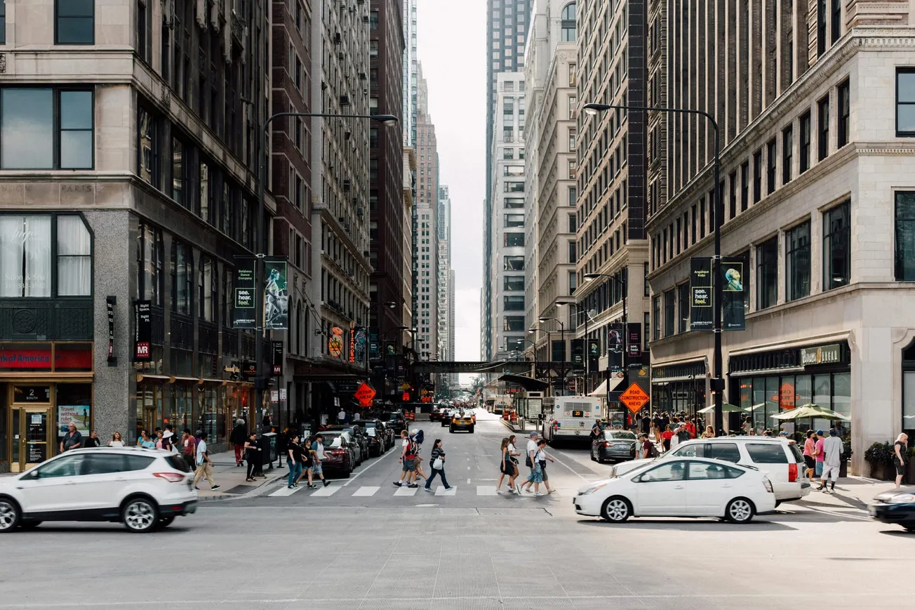 Busy city street lined with tall buildings, people crossing the crosswalk, cars waiting. The atmosphere is lively and bustling, typical of an urban setting.