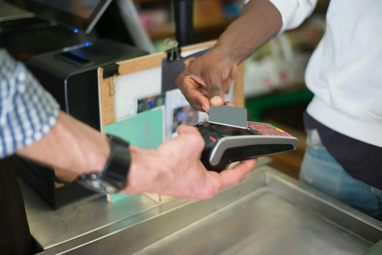 A person is using a credit card for a contactless payment on a card reader held by another person at a store checkout counter.