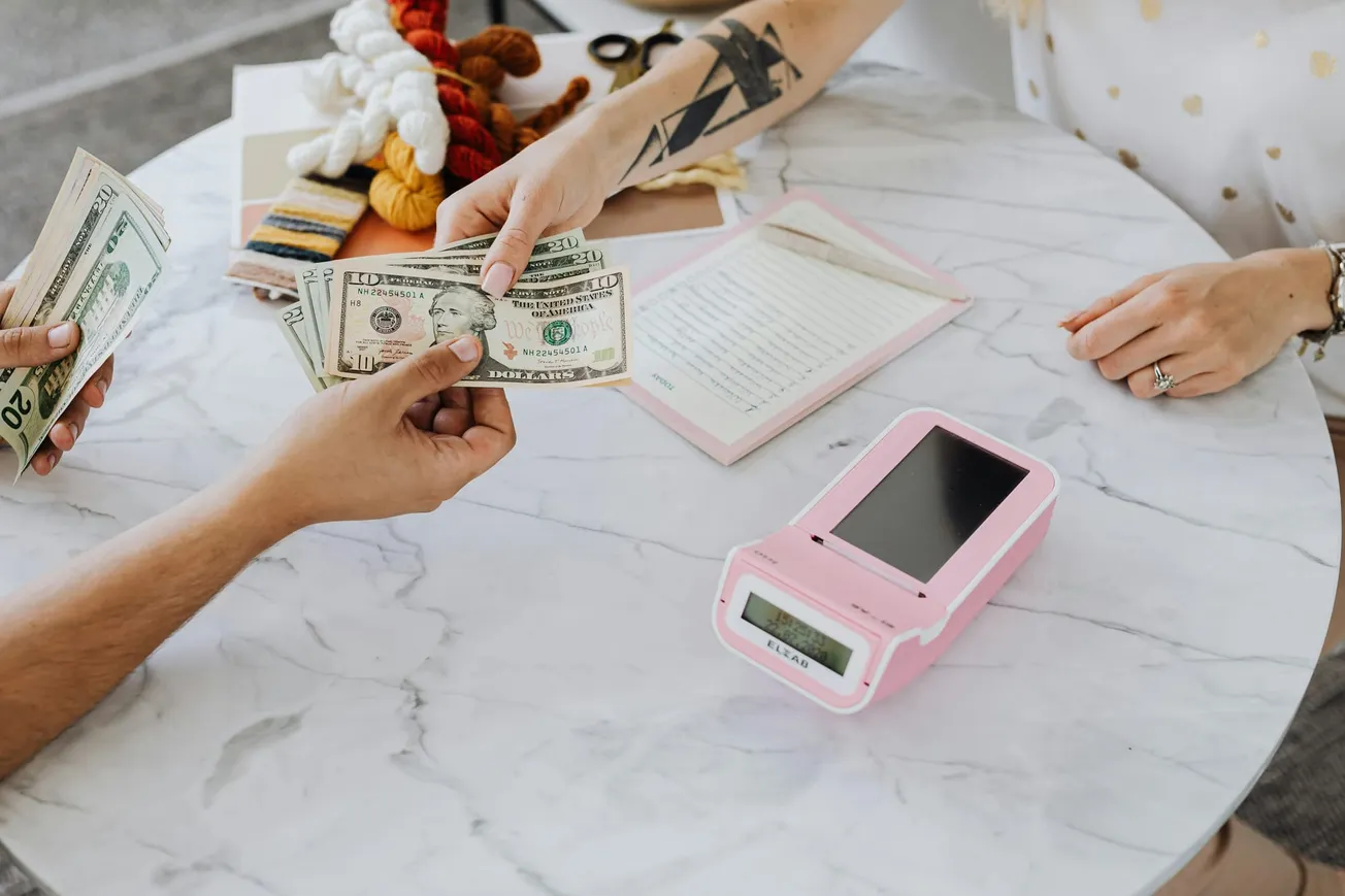A person with a geometric tattoo hands cash to another person across a marble table with a pink card machine. Yarn and a notepad are visible, suggesting a crafty or small business setting.