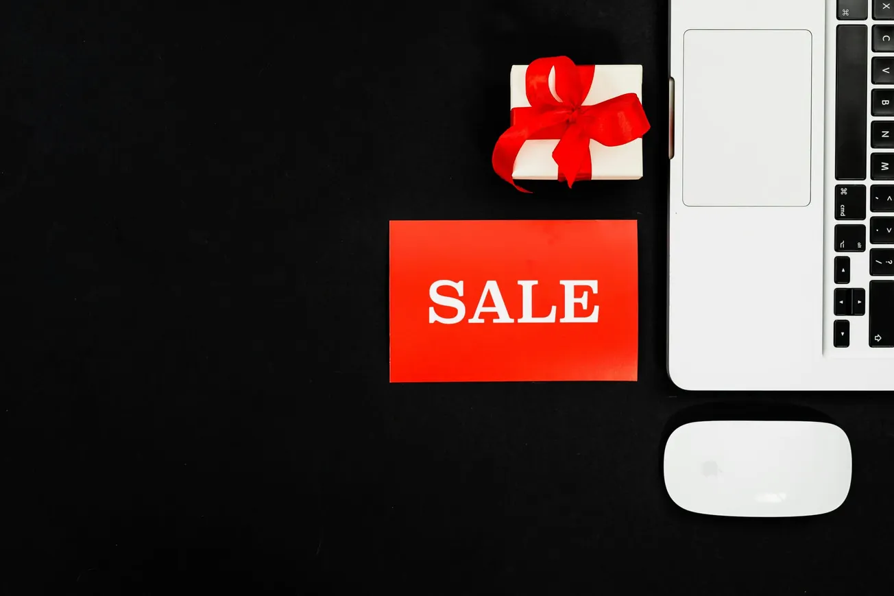 Alt text: "Image of a red sale card, a white gift box with a red bow, laptop corner, and wireless mouse on a black background, conveying a festive shopping theme."