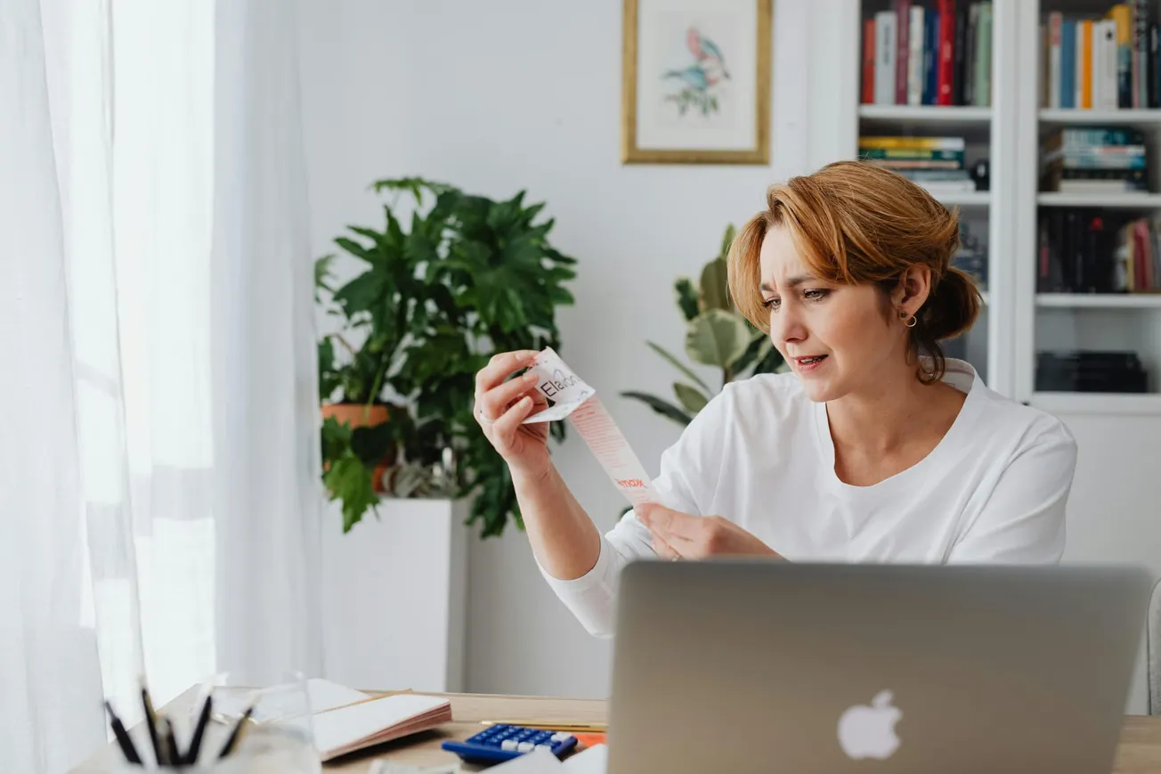 A woman in a white shirt sits at a desk, intently examining a receipt. Her expression is concerned. The room has a laptop, a calculator, and plants.