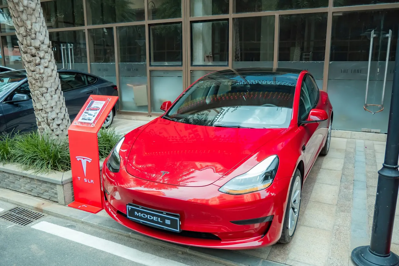 A red Tesla Model 3 parked on a city street beside a palm tree and a Tesla charging station. The background features a glass-fronted building.