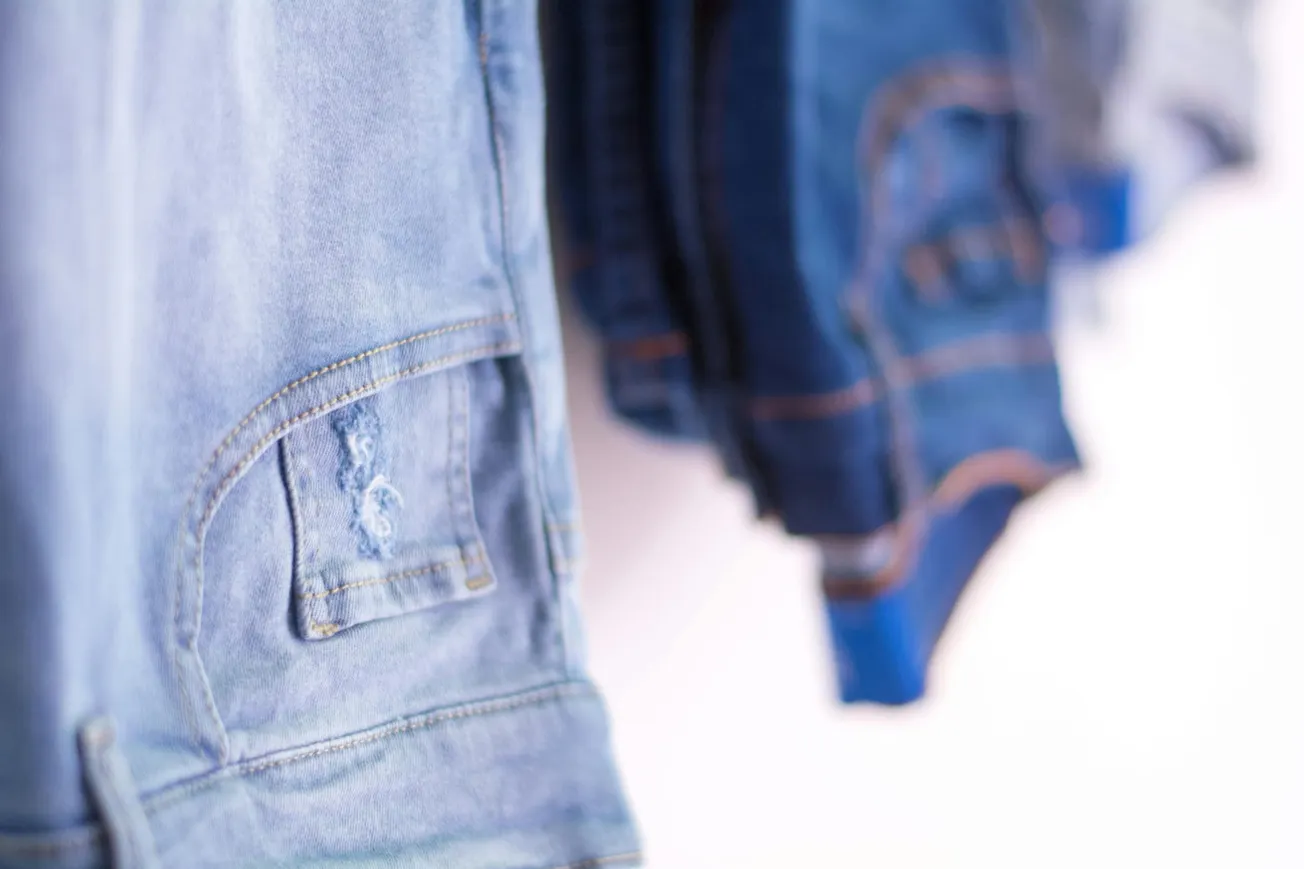 Close-up of faded blue denim jeans with a frayed pocket in focus, hanging alongside darker jeans. The image conveys a casual and worn look.