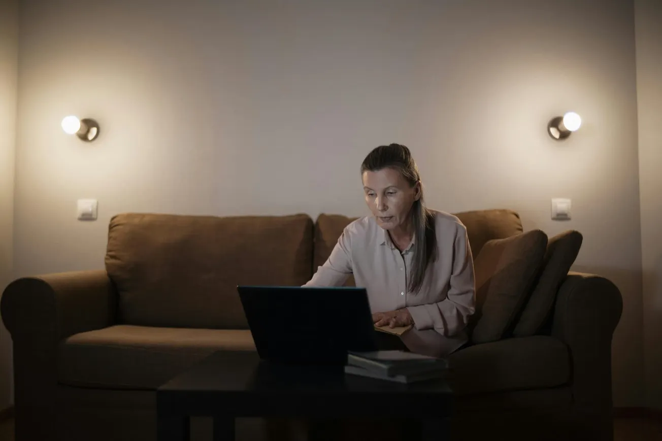 An older woman in a dimly lit room sits on a brown sofa, focused intently on her laptop. Two wall lights create a calm, serene ambiance.