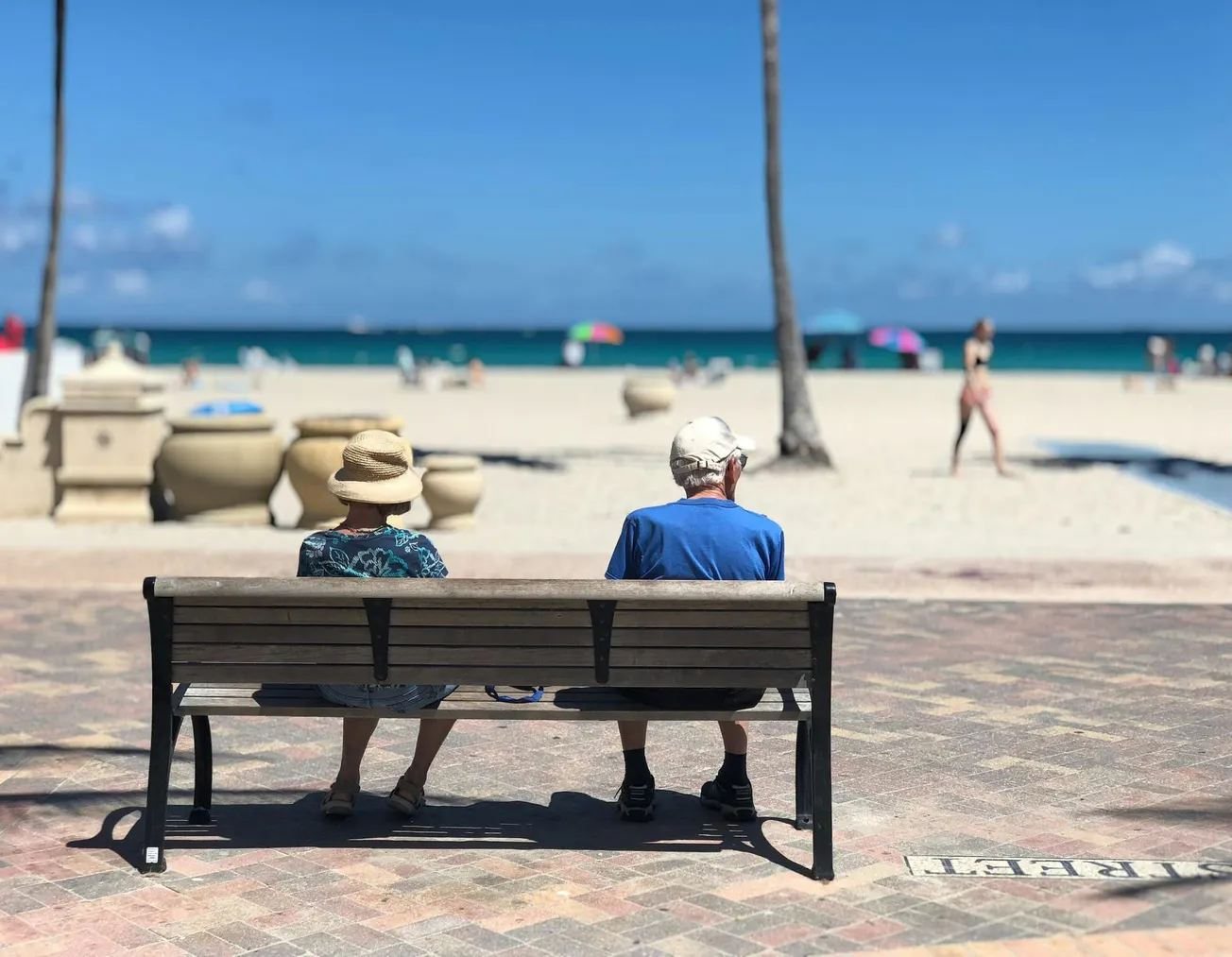 Elderly couple sitting on a bench by the beach, facing the ocean. Vibrant umbrellas dot the sandy shore, while a woman walks by. Peaceful, relaxed scene.