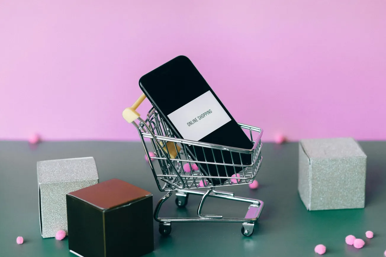 A smartphone displaying "Online Shopping" sits in a small shopping cart against a pink background. Nearby are three gift boxes.