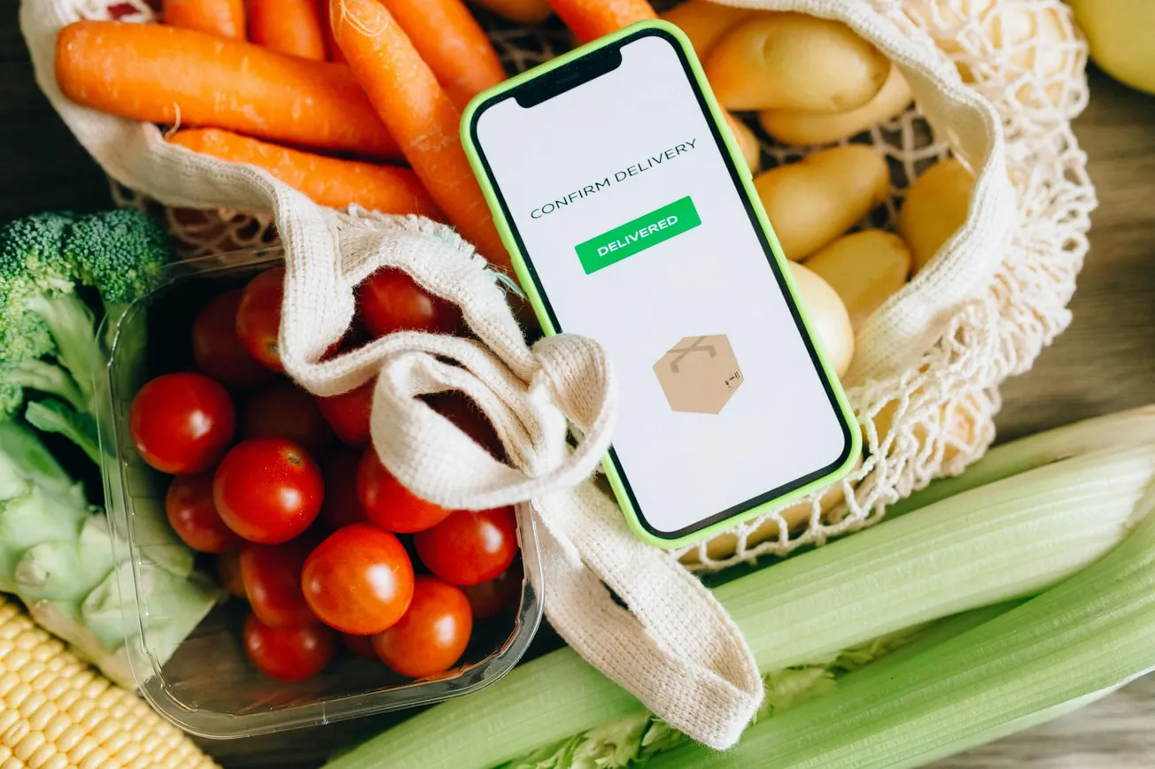 A smartphone with a "Delivered" screen rests among fresh vegetables, including carrots, tomatoes, broccoli, corn, and potatoes. The scene conveys freshness and convenience.