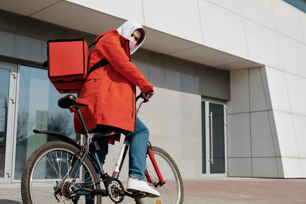 Delivery cyclist wearing a red jacket and white hoodie, carrying a red delivery bag on a bicycle. They are in front of a modern building.