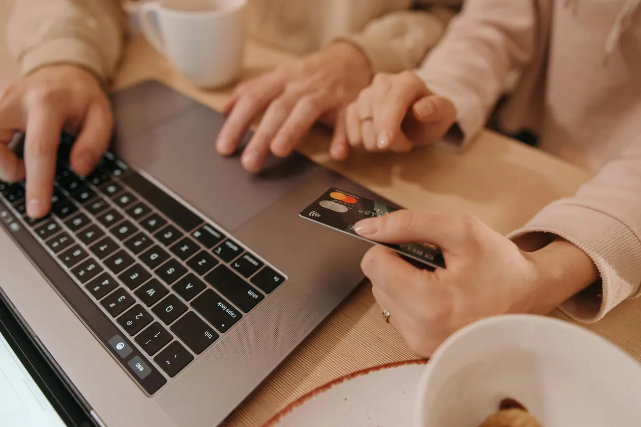 Two people use a laptop at a table. One holds a credit card, while the other types, suggesting online shopping. A coffee cup is nearby.